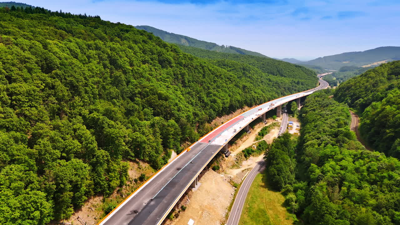Building of the freeway at the foot of green wooded mountains. Cars go by the road under the new highway. Aerial view