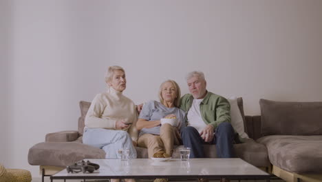 group of three senior people watching tv, talking and eating caramel popcorn while sitting on sofa at home 1