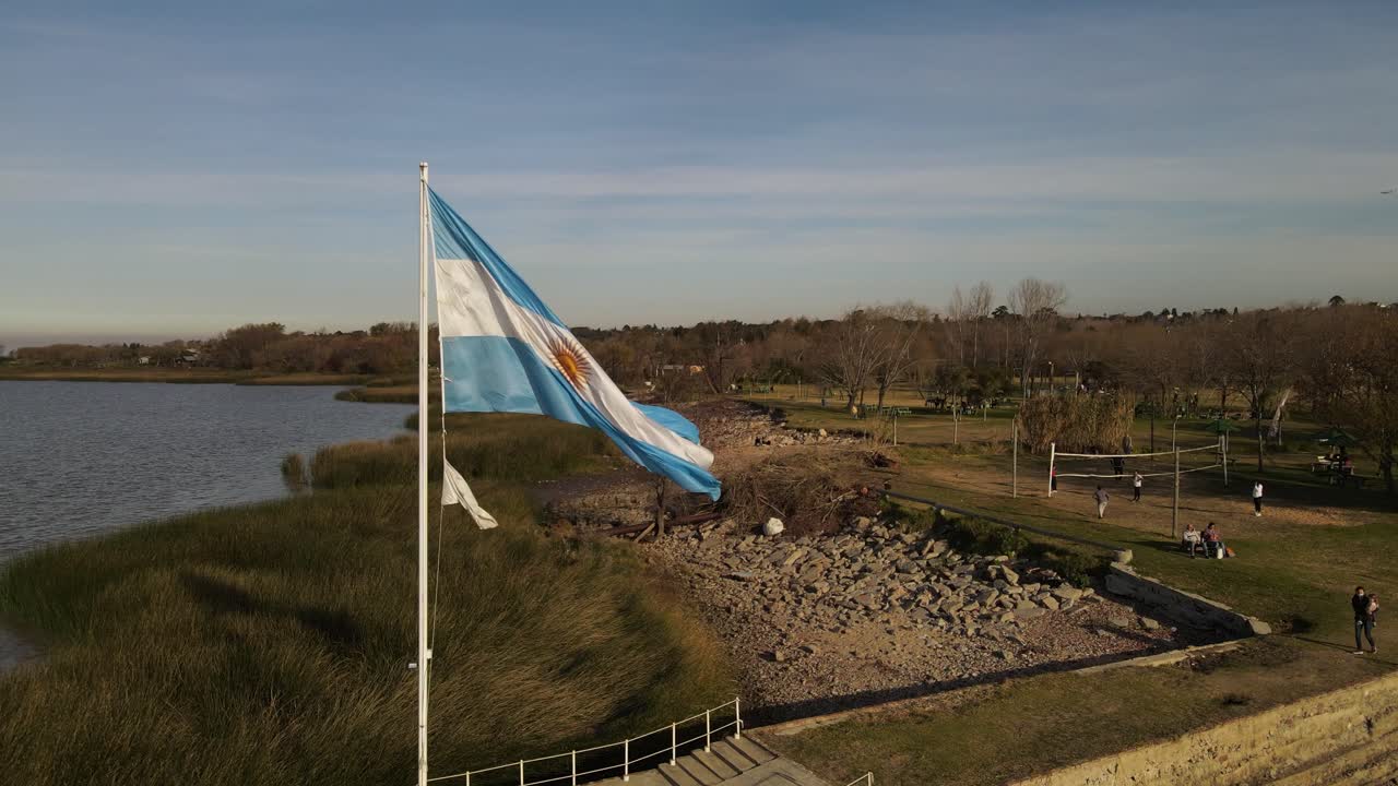 일몰 동안 바람에 흔들리는 아르헨티나 국기를 흔드는 슬로우 모션 트랙 샷 - 아름다운 공원과 해변을 배경으로