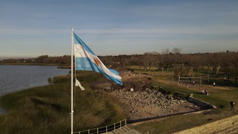 slow motion track shot of waving argentinian flag waving in wind during sunset - beautiful park and sea shore in background