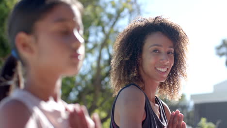 happy biracial mother practicing yoga meditation with daughter sitting in sunny garden, slow motion