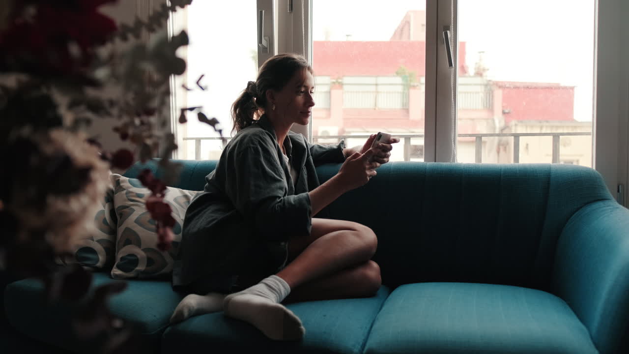 Giovane donna rilassata sul divano che usa lo smartphone vicino alla finestra