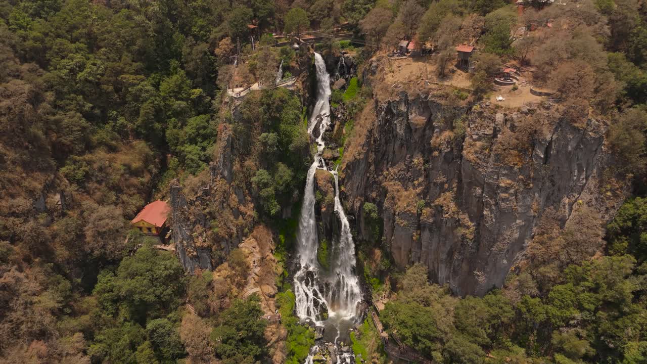 A breathtaking waterfall cascades down the green-brown cliffs of a mountain, with small houses nestled along the base, creating a serene and picturesque landscape