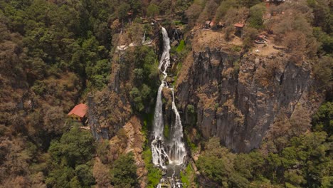 A breathtaking waterfall cascades down the green-brown cliffs of a mountain, with small houses nestled along the base, creating a serene and picturesque landscape