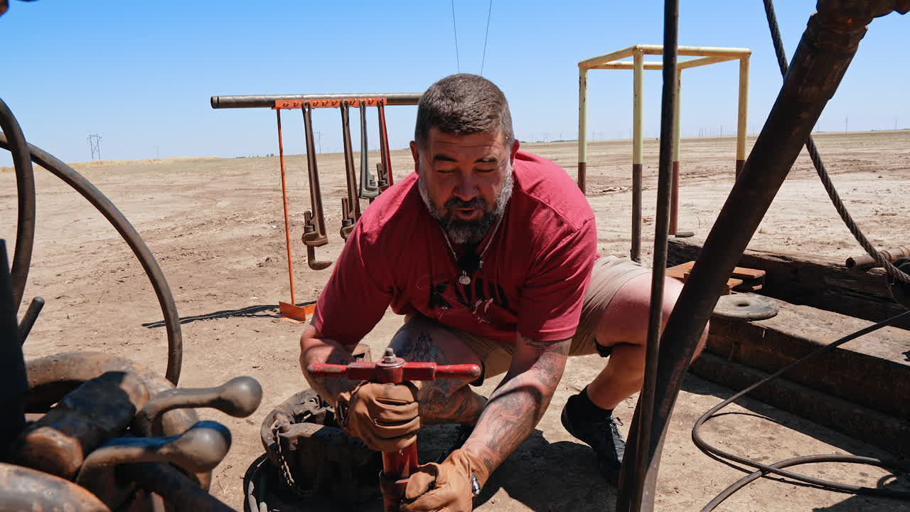 Talkative bearded man sits squatted adjusting the pipe with a faucet. Worker maintaining equipment at the gas producing site.