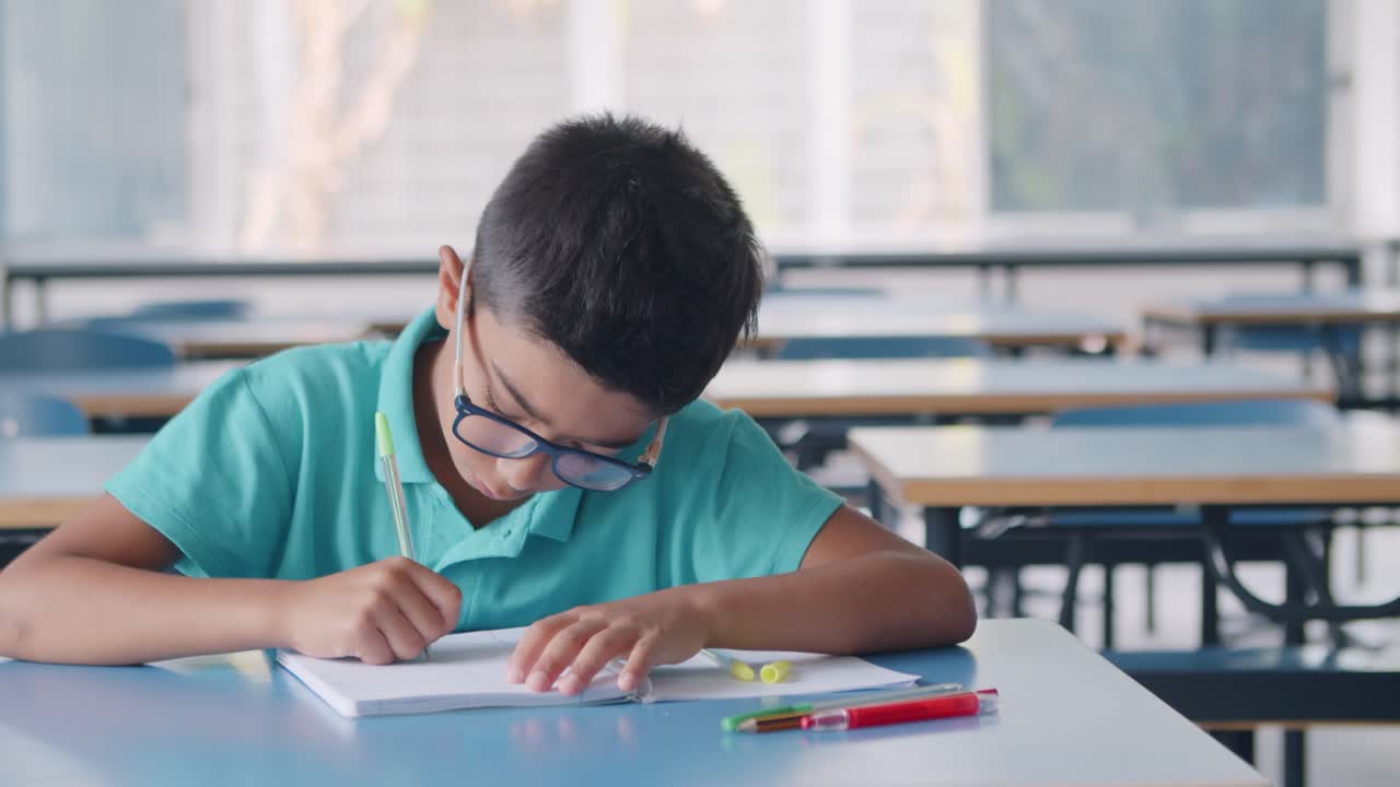 Niño alumno serio y aplicado con gafas escribiendo en un cuaderno en clase