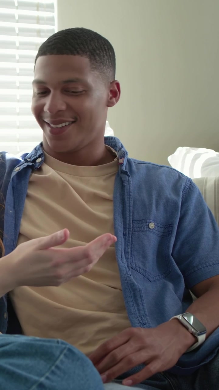 Video verticale: Uomo in camicia blu appoggiato al divano, sorridendo e reagendo ai gesti del partner.