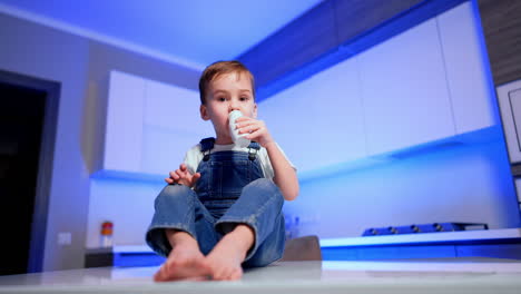 Adorable male kid in jeans romper sits on the table snacking. Cute boy drink yogurt from a bottle. Low angle view.