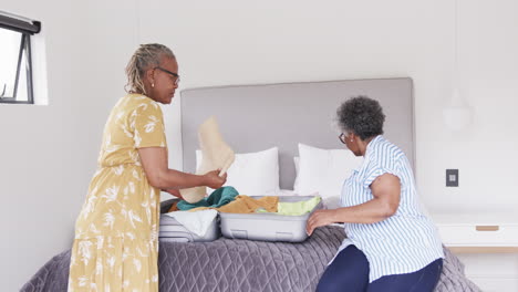 happy senior african american female friends unpacking suitcase on bed at holiday home, slow motion