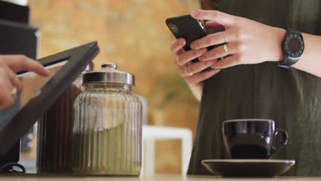 cliente caucásica que sostiene un teléfono inteligente y toma una taza de café del mostrador