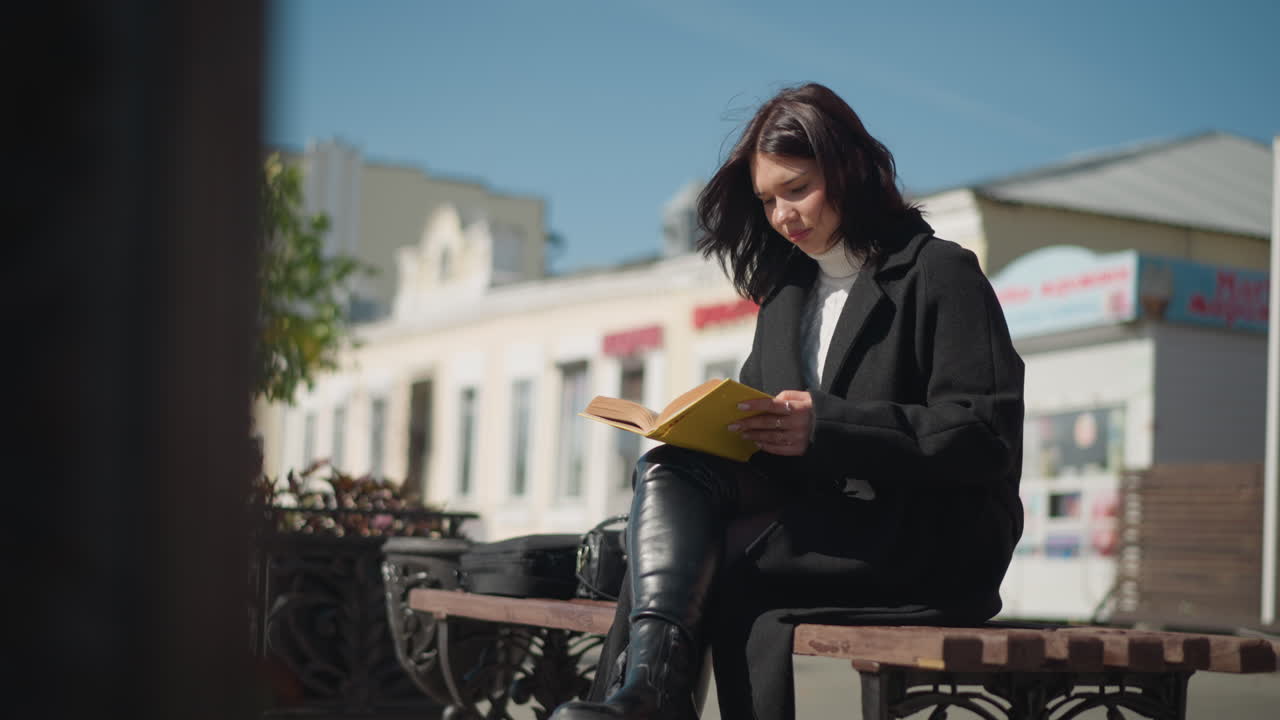 Woman sitting on a bench outdoors on a sunny day, focused on reading her book with yellow-edged pages flips to the next page, her hair blows gently in the breeze