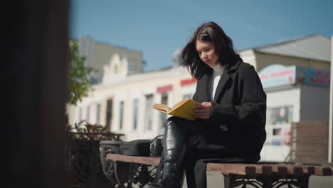 mujer sentada en un banco al aire libre en un día soleado, centrado en la lectura de su libro con páginas de borde amarillo voltea a la siguiente página, su cabello sopla suavemente en la brisa