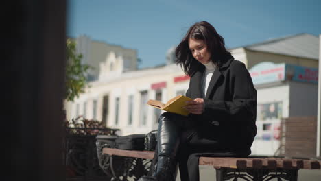 woman sitting on a bench outdoors on a sunny day, focused on reading her book with yellow-edged pages flips to the next page, her hair blows gently in the breeze