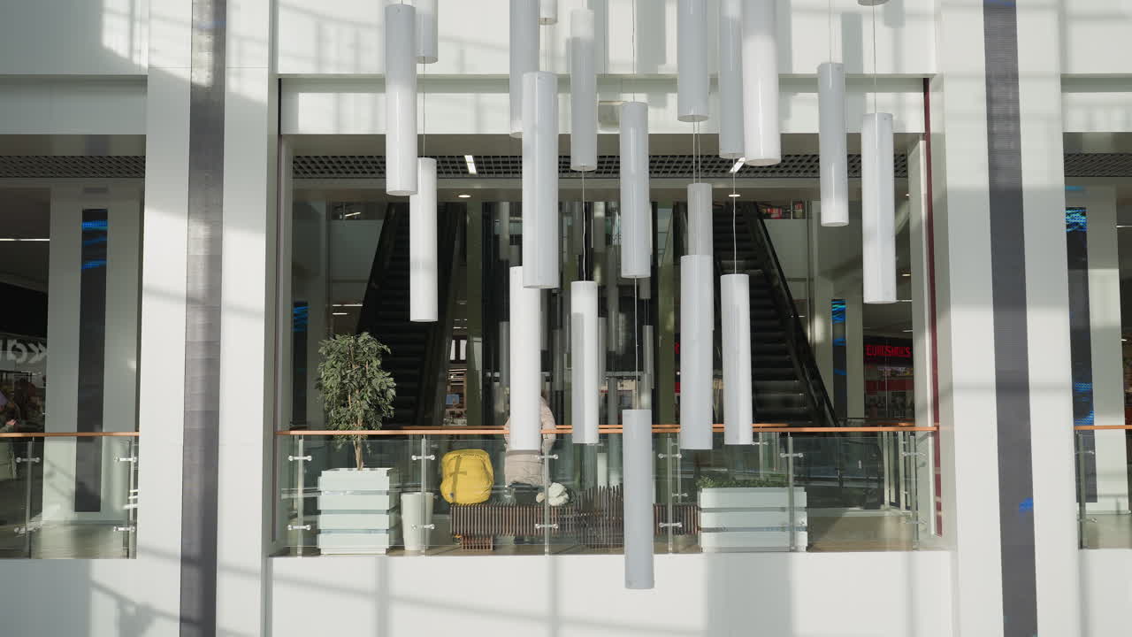 Wide view of stylish shopping mall interior featuring escalators, suspended white cylindrical light fixtures, potted plants, glass railings, and person rising from seat in sunlit open space