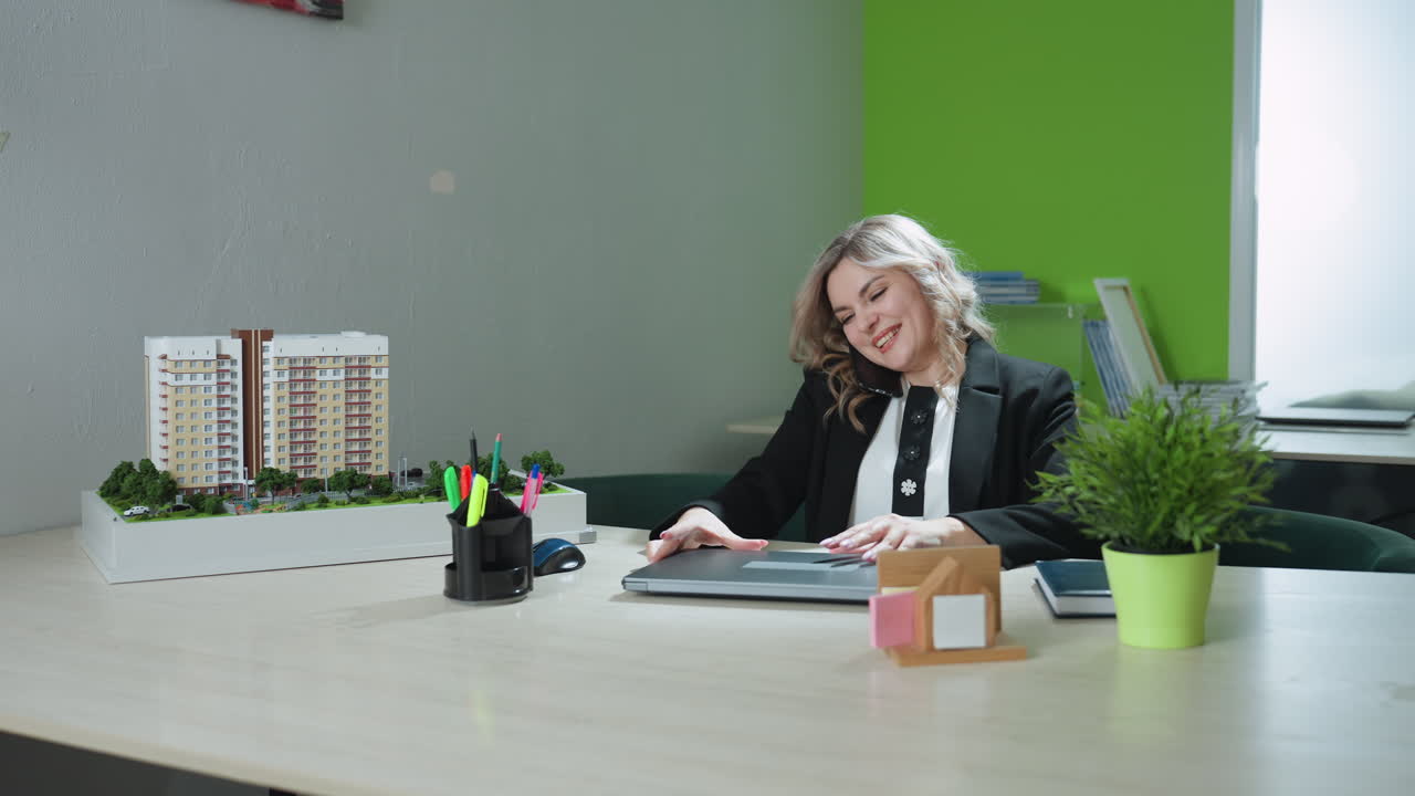 Woman seated in modern office opens laptop while speaking warmly on phone, surrounded by desk items including architectural model, potted plant, and organized files