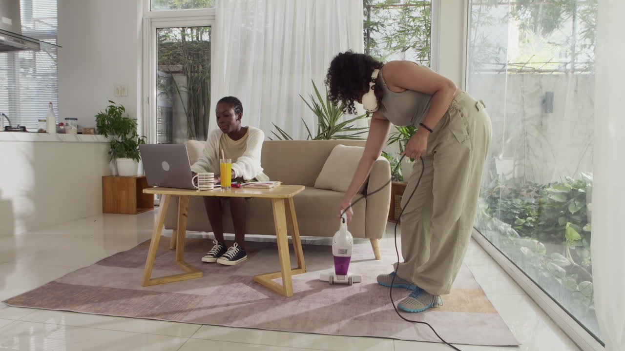 Girl Vacuum-cleaning while Another One Studying on Laptop