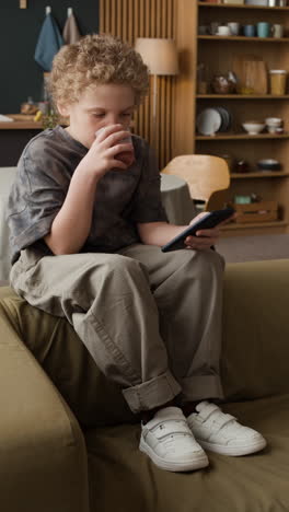 young boy sitting on a couch, drinking and using a smartphone
