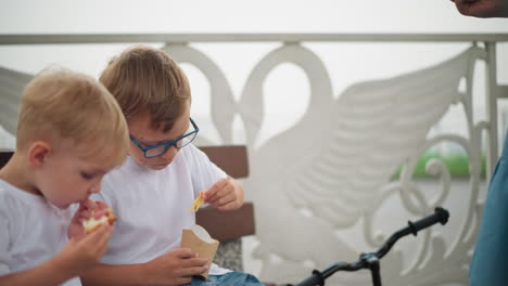 a mother is seen handing a bag of chips to her older son while the younger child is focused on eating a sausage roll