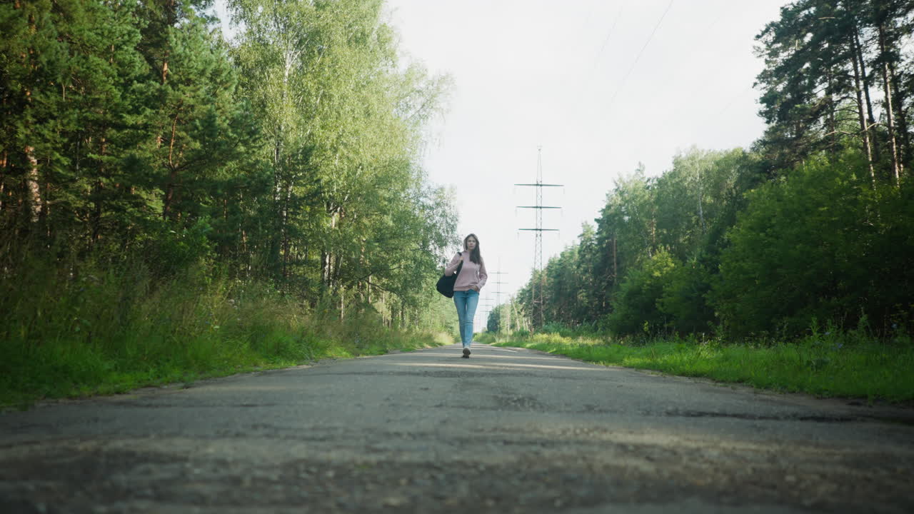 Young girl wearing pink hoodie and jeans walking along forest road with one hand in pocket and black bag over shoulder, surrounded by tall trees, green foliage, and power lines in background