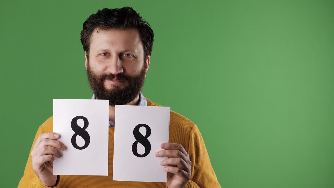 Man holding number 88 sign. Cheerful white bearded good-looking man in yellow sweater shows number eighty-eight on white sheet. Chrome key, green screen, copy space
