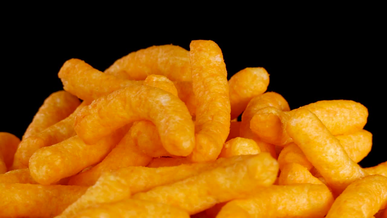 Cheese puffs rotate in detailed close-up, vibrant lighting, black background, macro food view