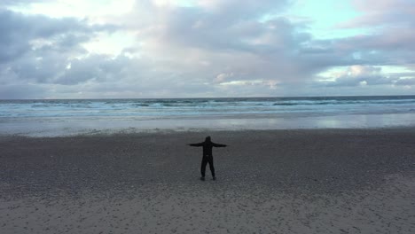 hombre de pie con los brazos abiertos disfrutando de la brisa del océano desde la playa de arena de playa de xago en asturias, españa