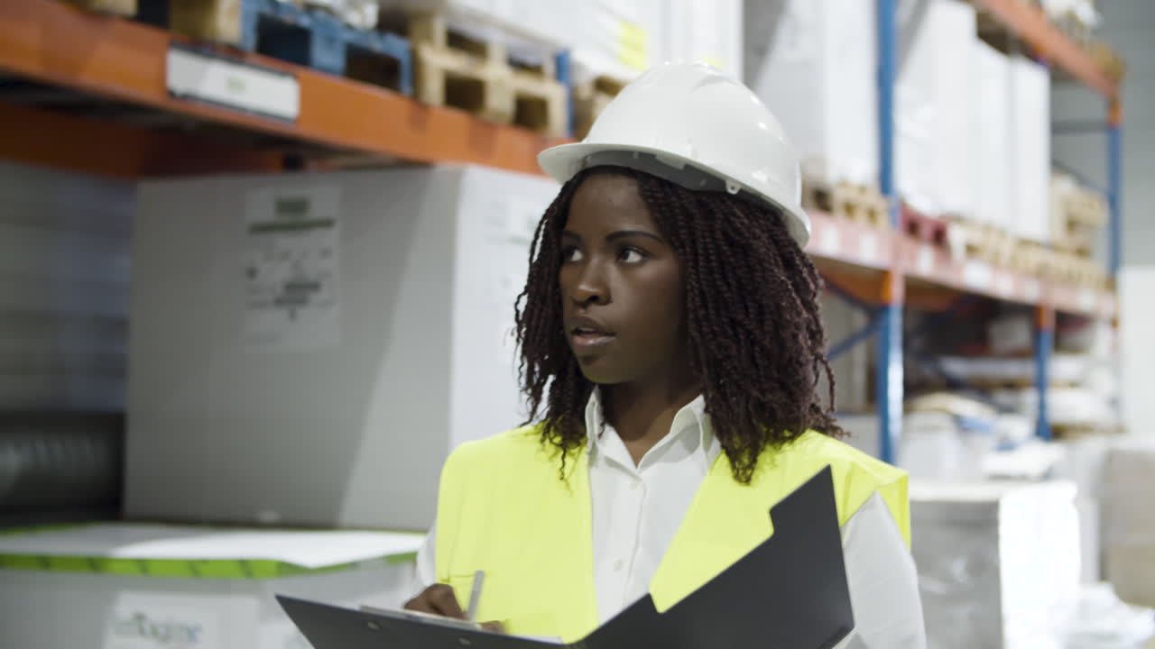 Serious African American female worker in helmet counting goods in stock