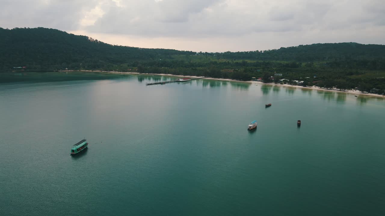Several tourist boats are moored in the quiet bay of Koh Rong Sanloem on a cloudy day as the water reflects off the trees near the calm. Wide drone lowering tilt shot