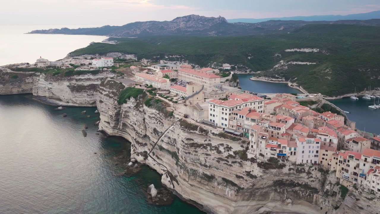 Una ripresa aerea al tramonto delle scogliere calcaree e degli edifici storici della città vecchia di Bonifacio in Corsica, Francia, con yacht di lusso nel porto protetto e un paesaggio scenografico di un'isola mediterranea