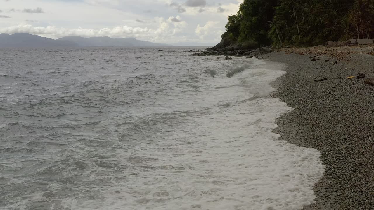dolly costera tomada a lo largo de la playa de guijarros con olas del océano con un fondo montañoso escénico en la bahía de looc, filipinas