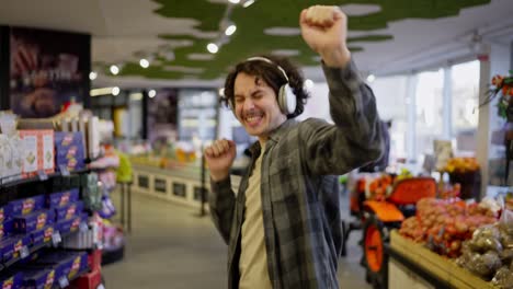 Cheerful-brunette-guy-in-a-checkered-shirt-wearing-white-headphones-listens-to-music-and-dances-in-a-grocery-store