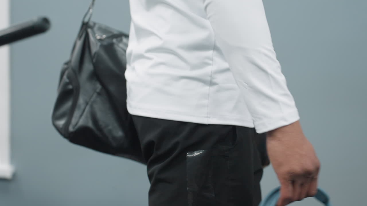 close up of man carrying black leather gym bag over shoulder and holding headphones as he drops bag onto bench inside bright fitness center corridor next to weight machine before workout begins