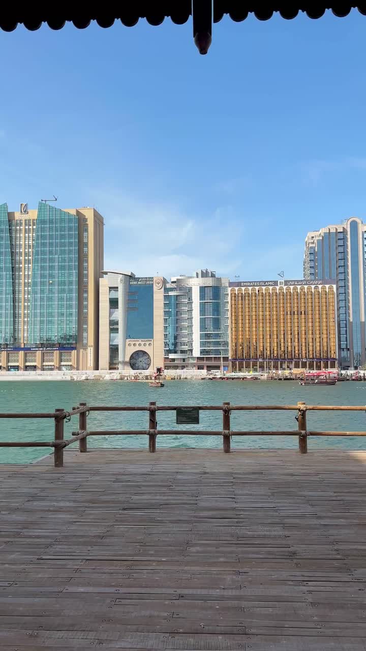 Vertical video of a tranquil waterfront deck overlooking modern glass high-rises including a unique circular-faced tower in Abu Dhabi. Ideal for mobile, social media, and stories