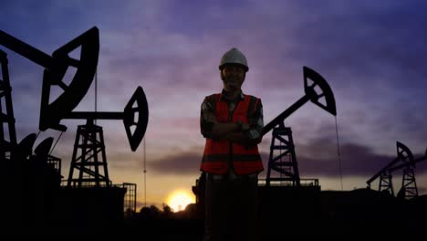 ingeniero asiático con casco de seguridad cruzando los brazos y sonriendo a la cámara mientras está de pie frente a las bombas de petróleo, durante la puesta o salida del sol