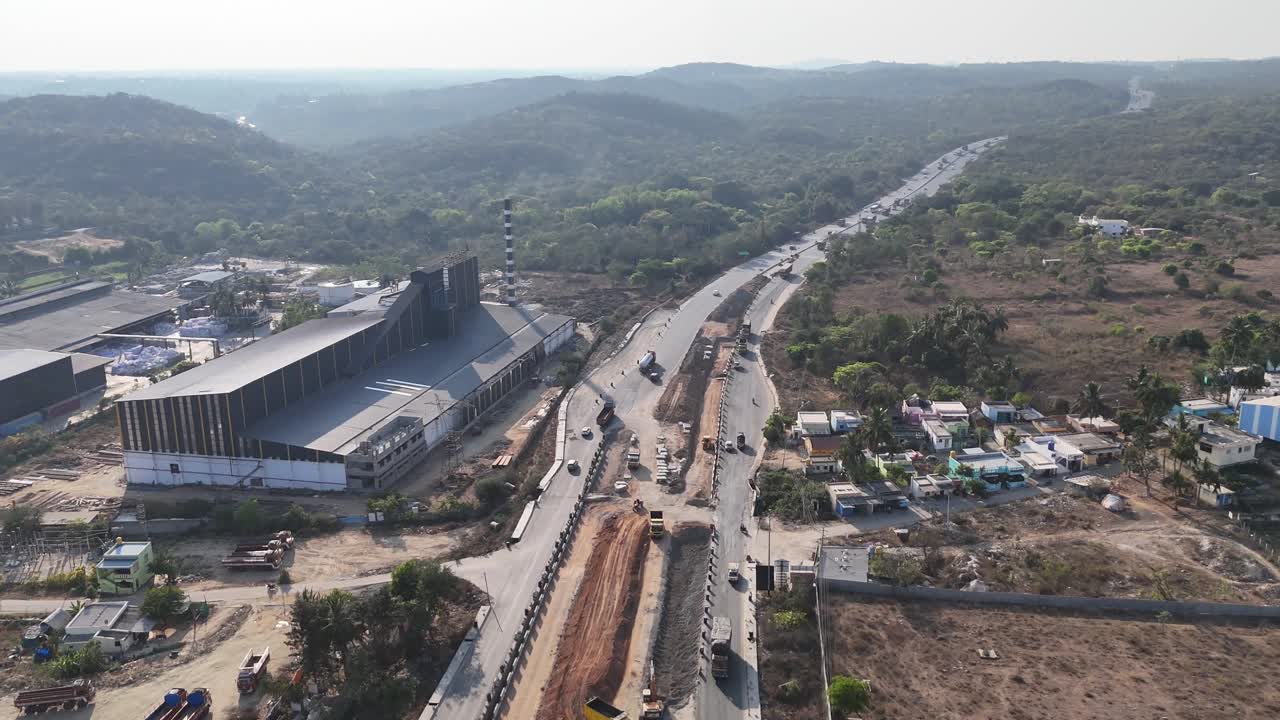 Aerial view of a highway under construction next to an active factory and a small town. Smoke mixing in the atmosphere, hills sky in the background. Industrial Expansion and Infrastructure Development
