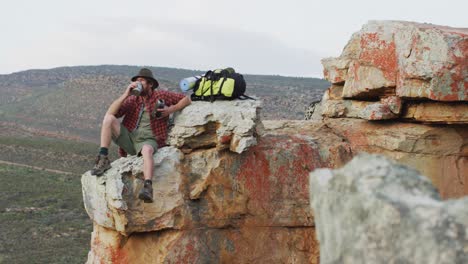 Superviviente-Masculino-Caucásico-Sentado-En-El-Pico-De-Una-Montaña-Rocosa-En-El-Desierto,-Descansando-Y-Bebiendo-Agua