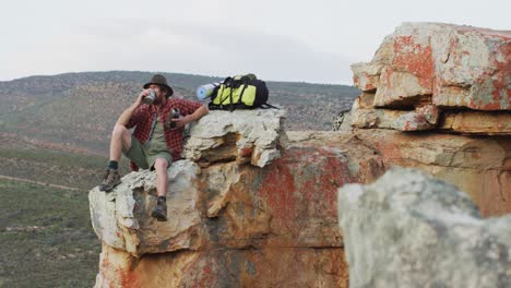 hombre caucásico sobreviviente sentado en el pico de una montaña rocosa en el desierto, descansando y bebiendo agua