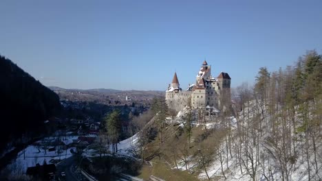 Aerial-Panorama-Of-Bran-Castle-On-A-Snowy-Hill-With-Blue-Sky-In-Background-In-Romania