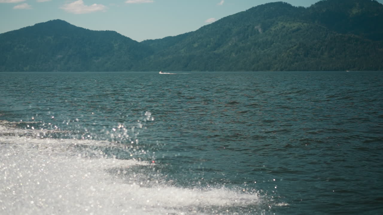 바물 위의 보트 트레일의 반이는 거품과 여름날에 큰 산의 그림자. 이국적인 리조트에서 경사 된 산맥에 대항하여 호수 표면에 요트 흔적