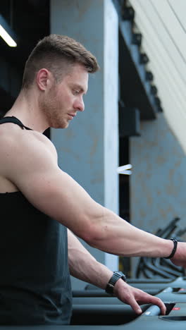 sportsman in fitness club. young handsome athlete switches on electronic treadmill in the gym. muscular man exercising on a simulator. profile view. vertical video