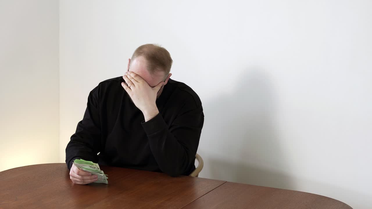 Uomo seduto al tavolo che conta denaro, si copre il viso per la frustrazione dopo aver capito che non è sufficiente. Concetto di debito, stress finanziario e difficoltà economiche.