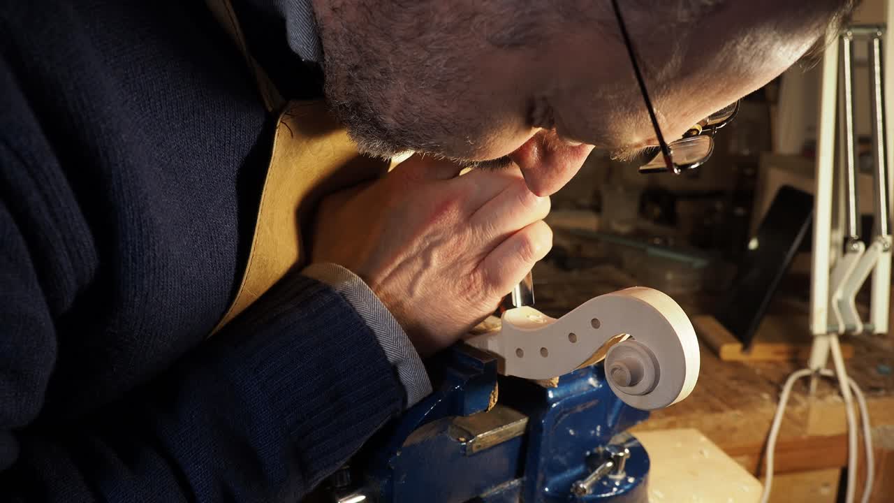 Focused artisan wearing glasses leans close to the violin scroll held in a vise, carefully carving the volute and pegbox with a chisel, applying precision, control, and traditional craftsmanship