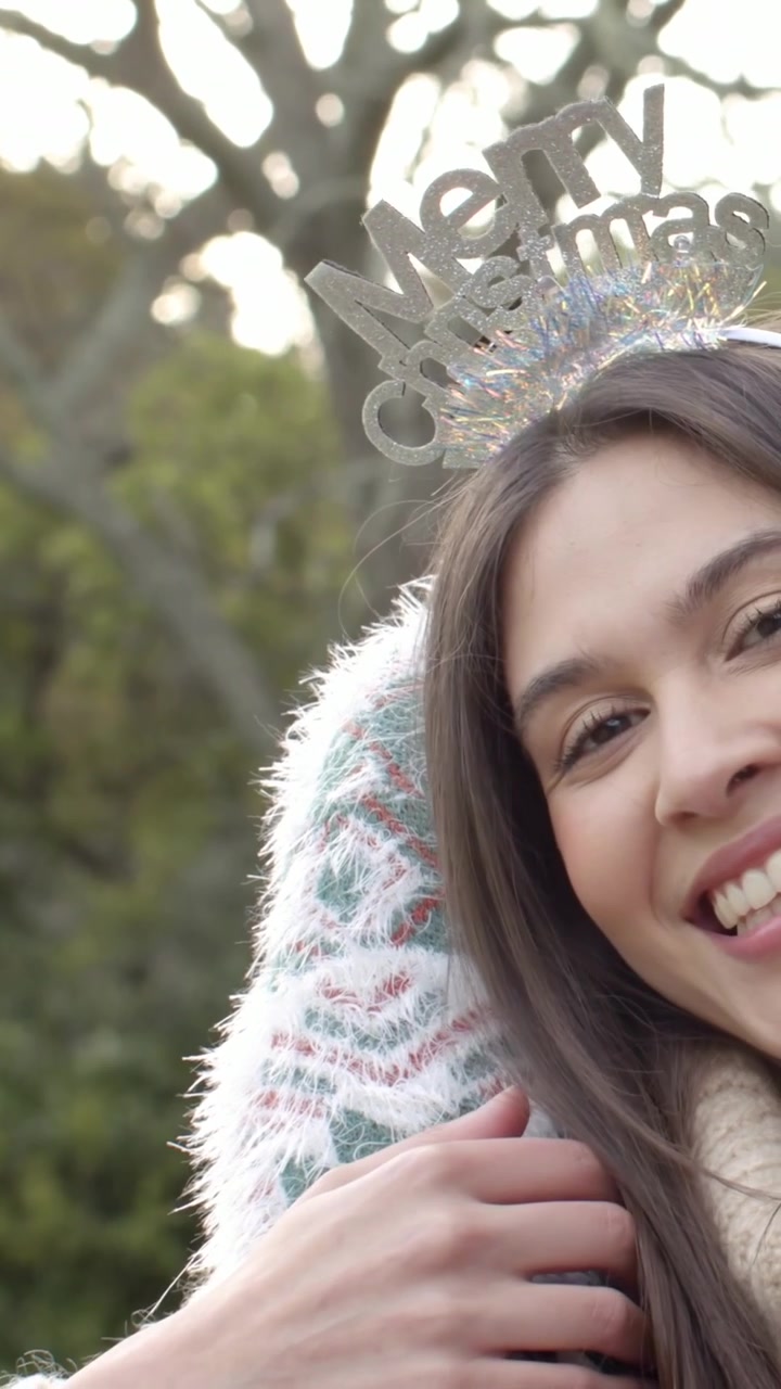 Video vertikal: Wanita berbusana rajut dengan bando "Merry Xmas" bersandar dan berpelukan di taman, merayakan.
