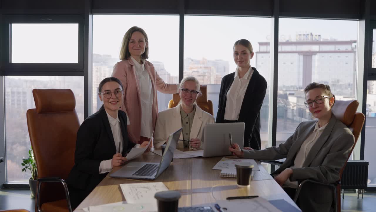 retrato de un grupo de chicas confiadas en trajes de negocios que llevan gafas en una mesa durante una reunión en una oficina moderna con grandes ventanas