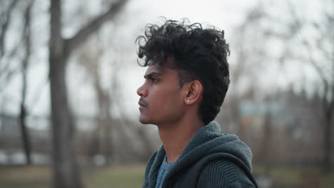 Black guy standing outdoors in park during early spring wearing gray hoodie, looking into distance with calm thoughtful expression, carrying black backpack, surrounded by blurred bare trees under cloudy