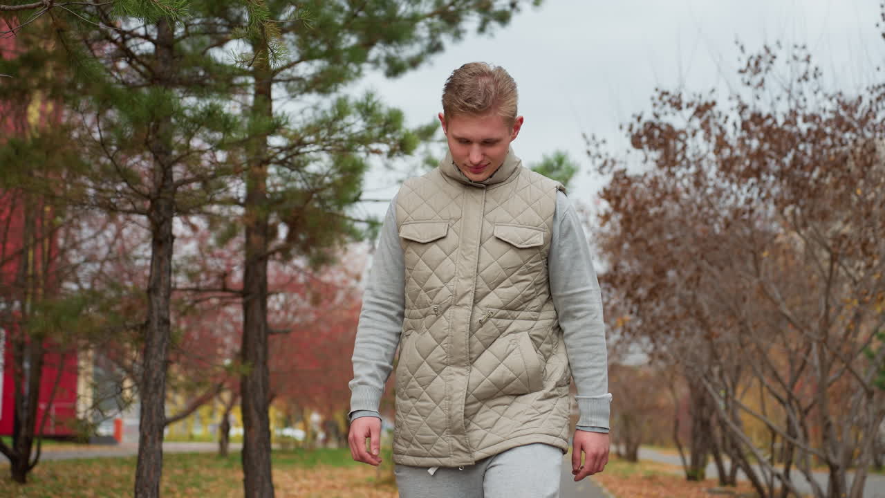 Young adult walking in cool breeze wearing jacket and joggers as pine trees sway with wind, autumn leaves cover ground, someone in red hoodie walks opposite direction in peaceful urban park setting