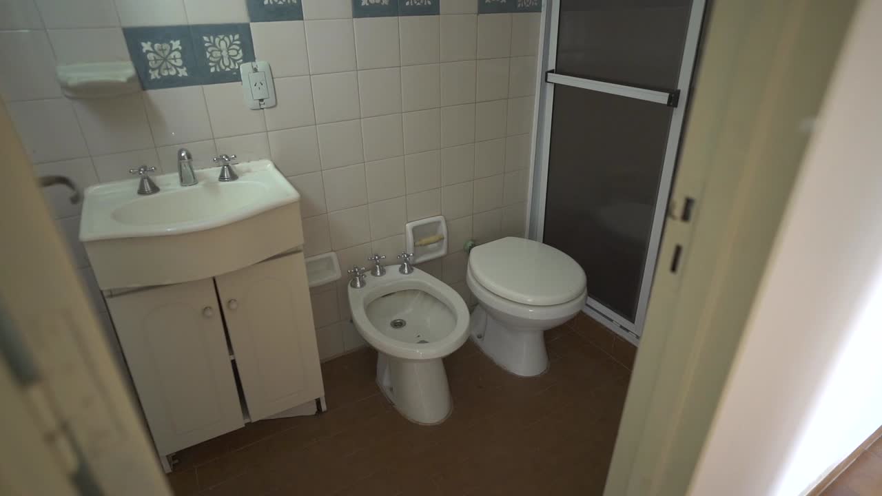 Smooth downward pan reveals modern bathroom layout, showcasing white sink, toilet with bidet, and glass shower enclosure. Perfect for rental listings.