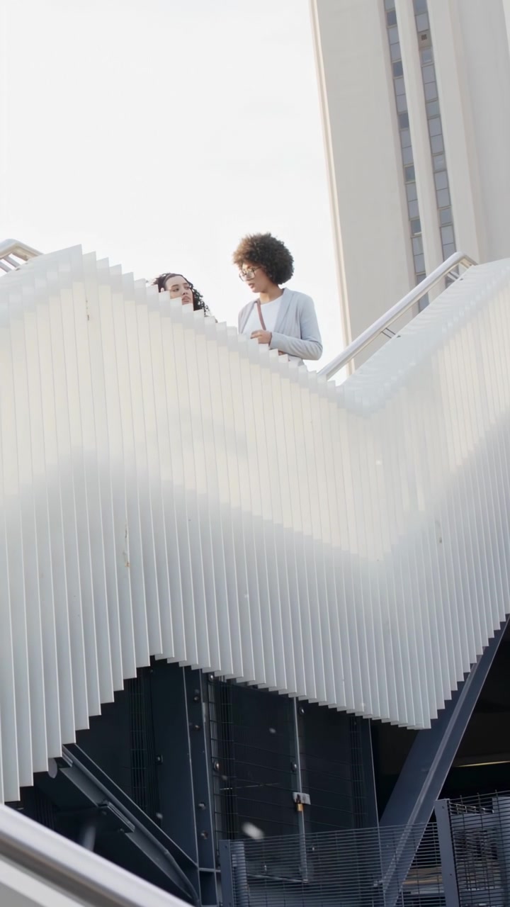 Video verticale: Due donne che camminano e parlano su un ponte urbano, con bagliori sulle costole e la conversazione che crea uno split visivo.