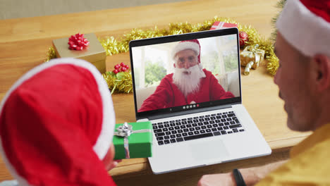 Padre-E-Hijo-Caucásicos-Con-Gorros-De-Papá-Noel-Usando-Una-Computadora-Portátil-Para-Una-Videollamada-Navideña-Con-Papá-Noel-En-La-Pantalla