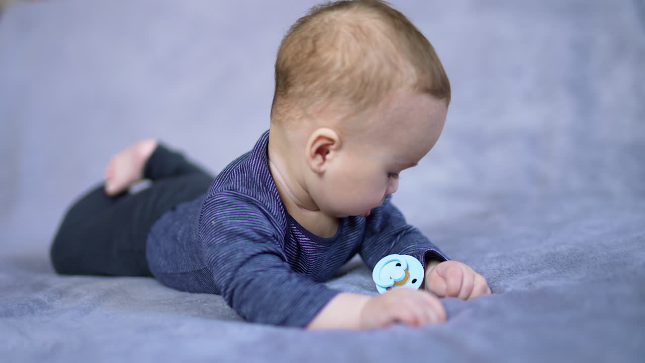Cute little boy in blue shirt lies on belly and stares at his hands. Caucasian kid on the bed with pacifier near him.